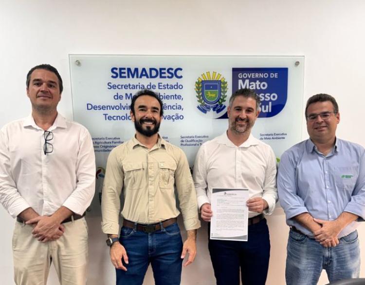 Quatro homens posam lado a lado em um ambiente institucional, em frente a uma placa da Secretaria de Estado de Meio Ambiente, Desenvolvimento, Ciência, Tecnologia e Inovação (SEMADESC) e do Governo de Mato Grosso do Sul. O terceiro homem, da esquerda para a direita, segura um documento. Todos estão vestidos com roupas sociais ou esporte fino e olham para a câmera durante o registro de uma reunião ou ato oficial. Ao fundo, aparecem os logotipos da SEMADESC e do Governo de Mato Grosso do Sul.